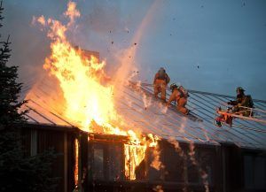 Firefighters on a sloped roof combating a raging house fire with tall flames and embers present.