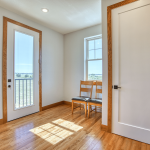 Interior doorway with white doors and oak trim, a window and balcony door visible, sunlight on hardwood floor, two wooden chairs nearby.