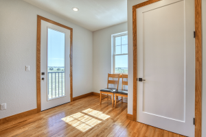 Interior doorway with white doors and oak trim, a window and balcony door visible, sunlight on hardwood floor, two wooden chairs nearby.