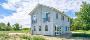 Two-story gray house with vertical siding, balconies, and large glass doors on a sunny day in a rural setting.