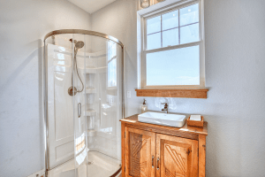 Shower stall with curved glass doors beside a wooden vanity and square sink under a window in a light gray bathroom.