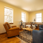 Cozy living room with yellow walls, three large windows, brown leather armchairs, a wooden coffee table, and a patterned rug.