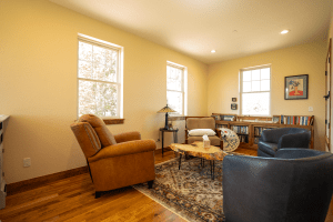Cozy living room with yellow walls, three large windows, brown leather armchairs, a wooden coffee table, and a patterned rug.