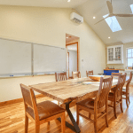 Bright open kitchen and dining area with a long wooden table and chairs, whiteboard on the wall, skylights, and a ceiling fan.