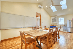 Bright open kitchen and dining area with a long wooden table and chairs, whiteboard on the wall, skylights, and a ceiling fan.