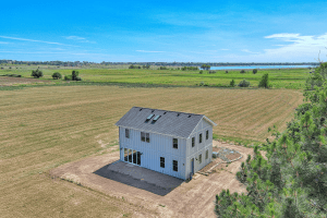 Two-story white house with a dark roof sits in a grassy rural field under a bright blue sky; a lake is visible in the distance,