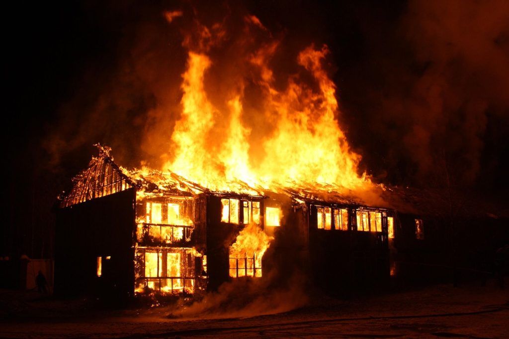 Two-story wooden house fully engulfed in flames at night with glowing windows
