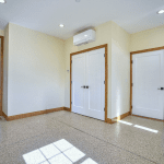 Hallway with double white doors under a wall-mounted air conditioner, beige walls, and speckled carpet; sunlight streams through windows in the adjacent rooms.