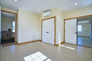 Hallway with double white doors under a wall-mounted air conditioner, beige walls, and speckled carpet; sunlight streams through windows in the adjacent rooms.
