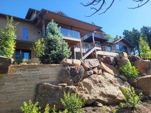 Modern multi-level house perched on a rocky hillside with wooden balconies and glass railings under a bright blue sky.