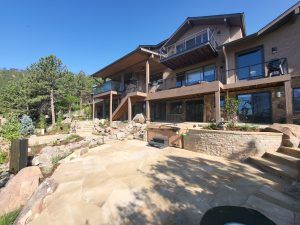 Modern multi‑level house with wooden decks and glass railings overlooking rocky landscaping and clear blue sky.