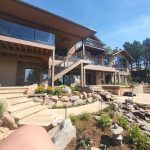 Modern multi-level house with wood and stone facade, glass balconies, and a rocky landscaped yard under a blue sky.