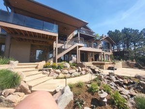 Modern multi-level house with wood and stone facade, glass balconies, and a rocky landscaped yard under a blue sky.