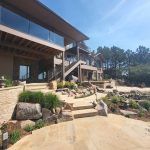 Modern multi‑level residential building with wooden beams, glass railings, and stone terrace under a clear blue sky.