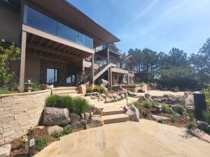 Modern multi‑level residential building with wooden beams, glass railings, and stone terrace under a clear blue sky.