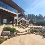 Modern multi-level building with glass balconies, stone walls, and rocky landscaped steps under a clear blue sky