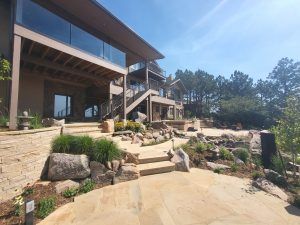 Modern multi-level building with glass balconies, stone walls, and rocky landscaped steps under a clear blue sky