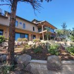 Multi-story beige townhouse with a wooden deck and glass railing, surrounded by a rock garden and shrubs, under a clear blue sky.