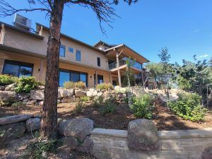 Multi-story beige townhouse with a wooden deck and glass railing, surrounded by a rock garden and shrubs, under a clear blue sky.