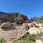 Residential building on a rocky hillside with a stone retaining wall and greenery, under a clear blue sky.