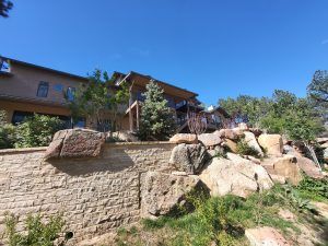 Residential building on a rocky hillside with a stone retaining wall and greenery, under a clear blue sky.