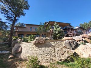 Residential building on a rocky hillside with a stone retaining wall and evergreen trees under a clear blue sky