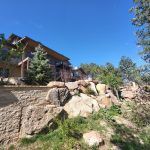 Residential building with wooden balconies on a rocky hillside, blue sky overhead, and stone retaining wall in the foreground surrounded by trees.