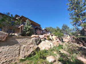 Residential building with wooden balconies on a rocky hillside, blue sky overhead, and stone retaining wall in the foreground surrounded by trees.