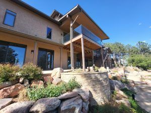 Modern multi-level house with an outdoor wooden balcony, glass railing, and a stone-terraced rock garden under a clear blue sky.