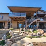 Front view of a multi-level backyard patio with wooden decks, glass railing, and stairs against a clear blue sky.