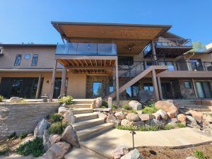 Front view of a multi-level backyard patio with wooden decks, glass railing, and stairs against a clear blue sky.