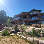 Modern hillside house with multiple balconies and glass railings, surrounded by a rock garden and drought-tolerant plants under a bright blue sky.
