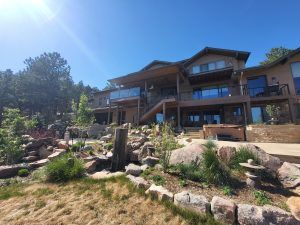 Modern hillside house with multiple balconies and glass railings, surrounded by a rock garden and drought-tolerant plants under a bright blue sky.