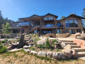 Modern two-story house with large glass windows, wooden decks, and metal railings overlooking a rocky, landscaped yard on a sunny day.