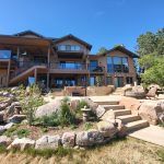 Modern multi-level house with a wooden deck, large glass windows, and stone foundation, set against a bright blue sky with rocky, landscaped foreground.