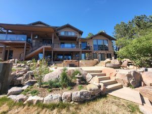 Modern multi-level house with a wooden deck, large glass windows, and stone foundation, set against a bright blue sky with rocky, landscaped foreground.