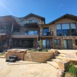 Modern two-story house with stone base, large windows, and multiple balconies under a clear blue sky.
