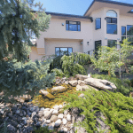 Modern beige, two-story house with large windows, set in a landscaped yard with a rocky stream and greenery in the foreground.