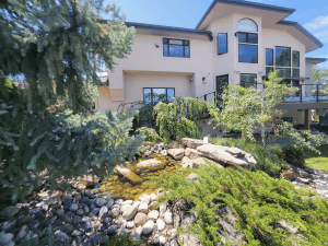 Modern beige, two-story house with large windows, set in a landscaped yard with a rocky stream and greenery in the foreground.