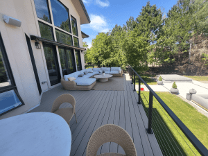 Outdoor balcony with a beige sectional sofa, wicker chairs, and a round coffee table overlooking a green lawn and trees beyond the railing, in front of a modern house with large windows.
