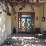 Wooden double front door with decorative glass panels under a covered porch, purple leaves overhead casting shadows on beige walls; outdoor seating on the right.