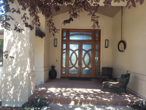 Wooden double front door with decorative glass panels under a covered porch, purple leaves overhead casting shadows on beige walls; outdoor seating on the right.