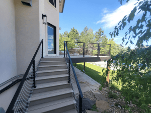 Outdoor staircase along a modern home with metal cable railings, leading to a deck with a patio table under a blue sky.