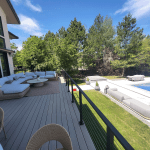 Sunlit backyard deck with a large white sectional sofa and wicker chairs, overlooking a rectangular pool and green lawn under a blue sky