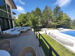 Sunlit backyard deck with a large white sectional sofa and wicker chairs, overlooking a rectangular pool and green lawn under a blue sky