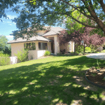 Suburban front yard with a beige single-story house, tiled roof, and large shade trees under a bright blue sky.