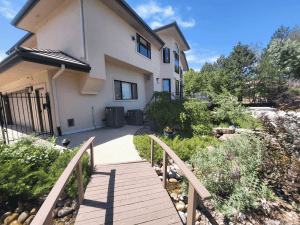 Two-story beige house with large windows, viewed from a wooden bridge over a rocky, landscaped yard on a sunny day.