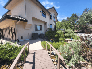 Wooden walkway with railings leading to a beige two-story house, surrounded by shrubs and a rock garden on a sunny day in clear weather.