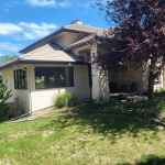 Front view of a beige house with a tiled sloped roof, large windows, and a shaded porch, set in a green yard under a blue sky with trees nearby.