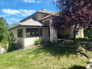 Front view of a beige house with a tiled sloped roof, large windows, and a shaded porch, set in a green yard under a blue sky with trees nearby.